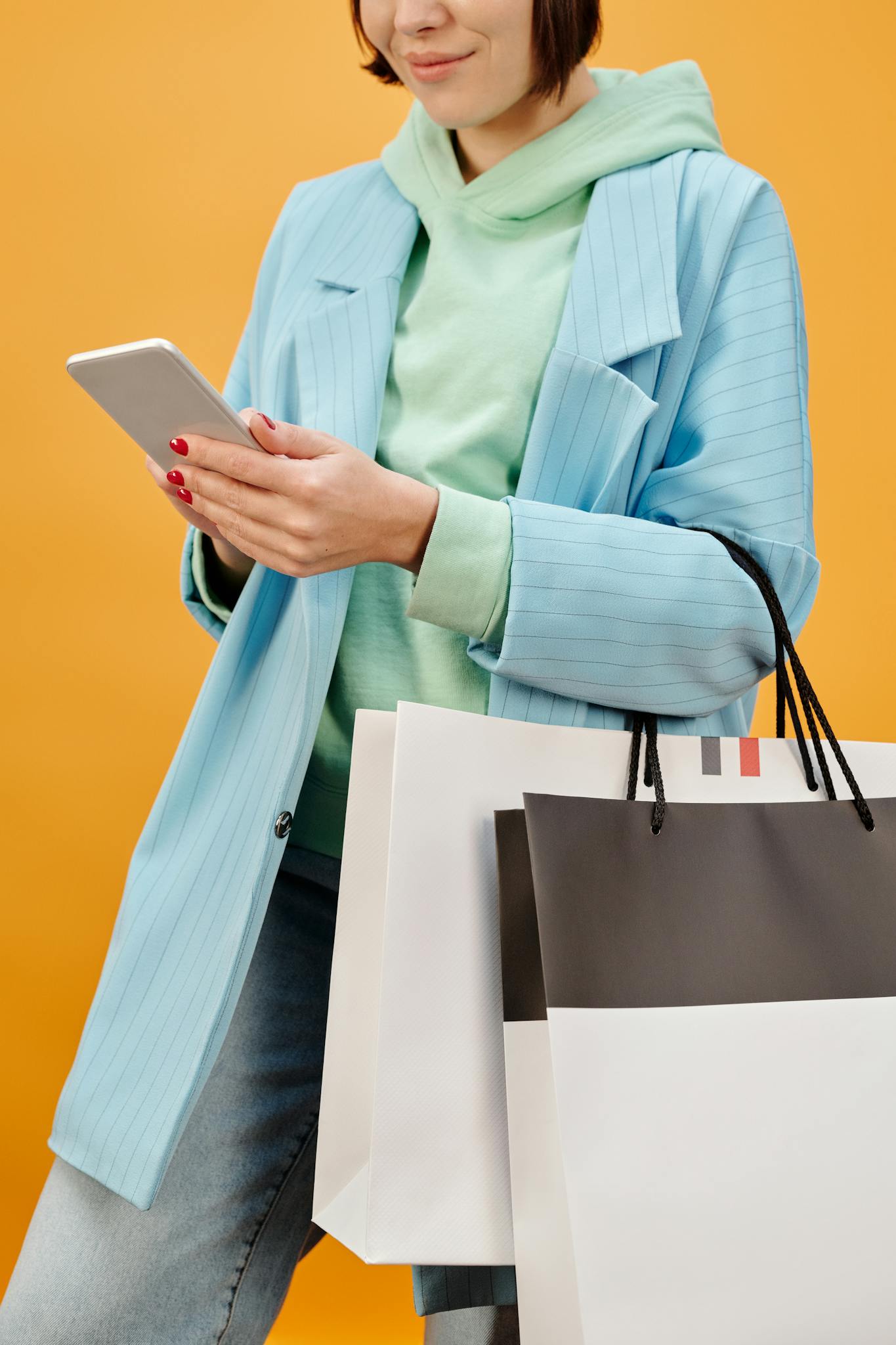 A fashionable woman in blue coat holding shopping bags and checking her phone against a yellow background.