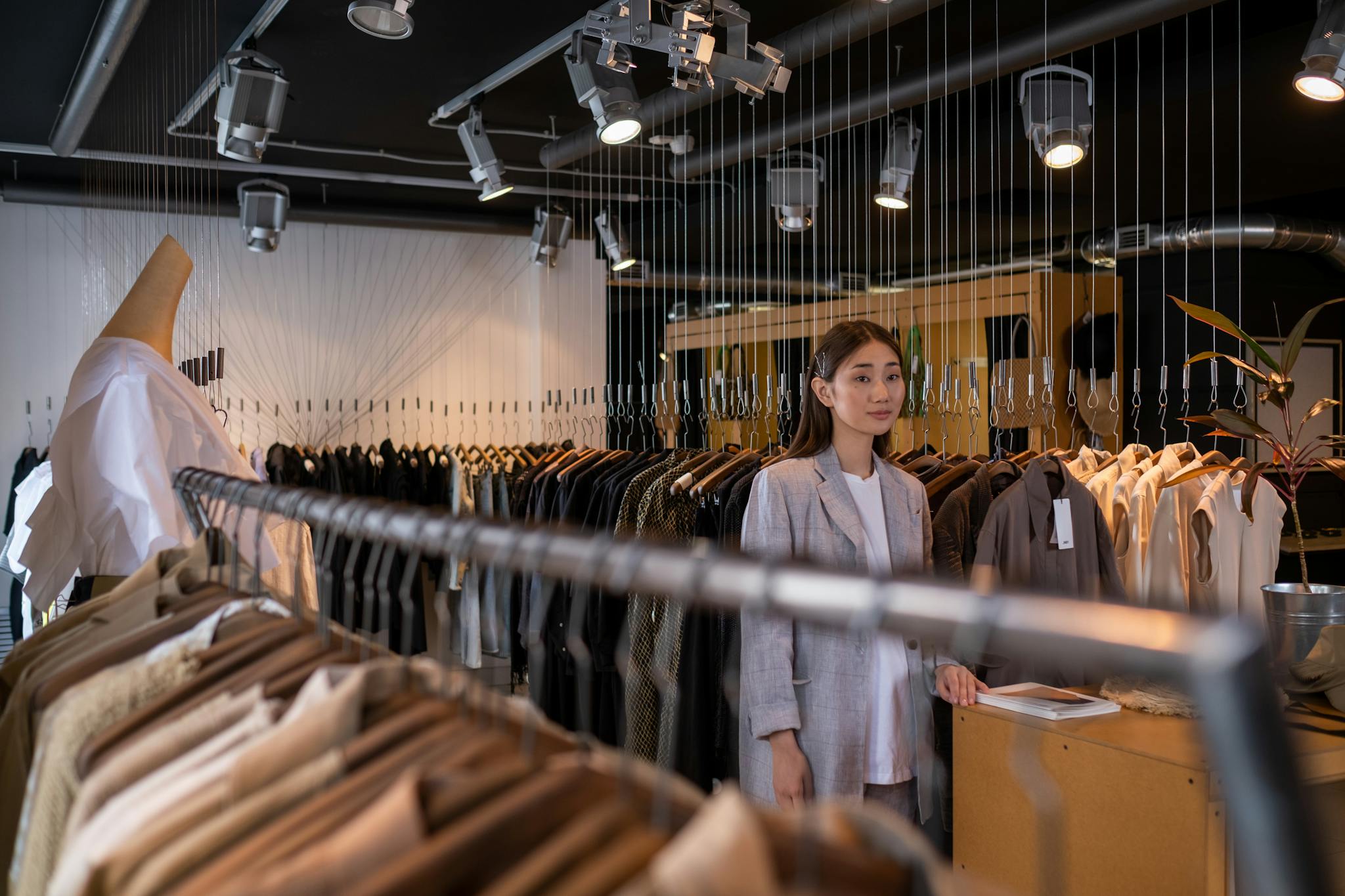 A stylish Asian woman stands confidently in a modern boutique surrounded by chic clothing options.