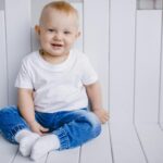Charming portrait of a smiling baby boy in a white t-shirt and blue jeans, sitting indoors.