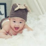 A joyful baby wearing a brown knitted hat lies on a soft surface, radiating happiness.