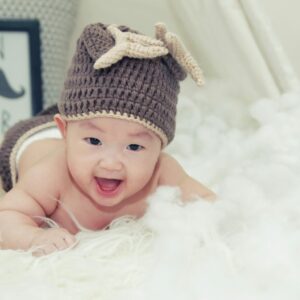 A joyful baby wearing a brown knitted hat lies on a soft surface, radiating happiness.