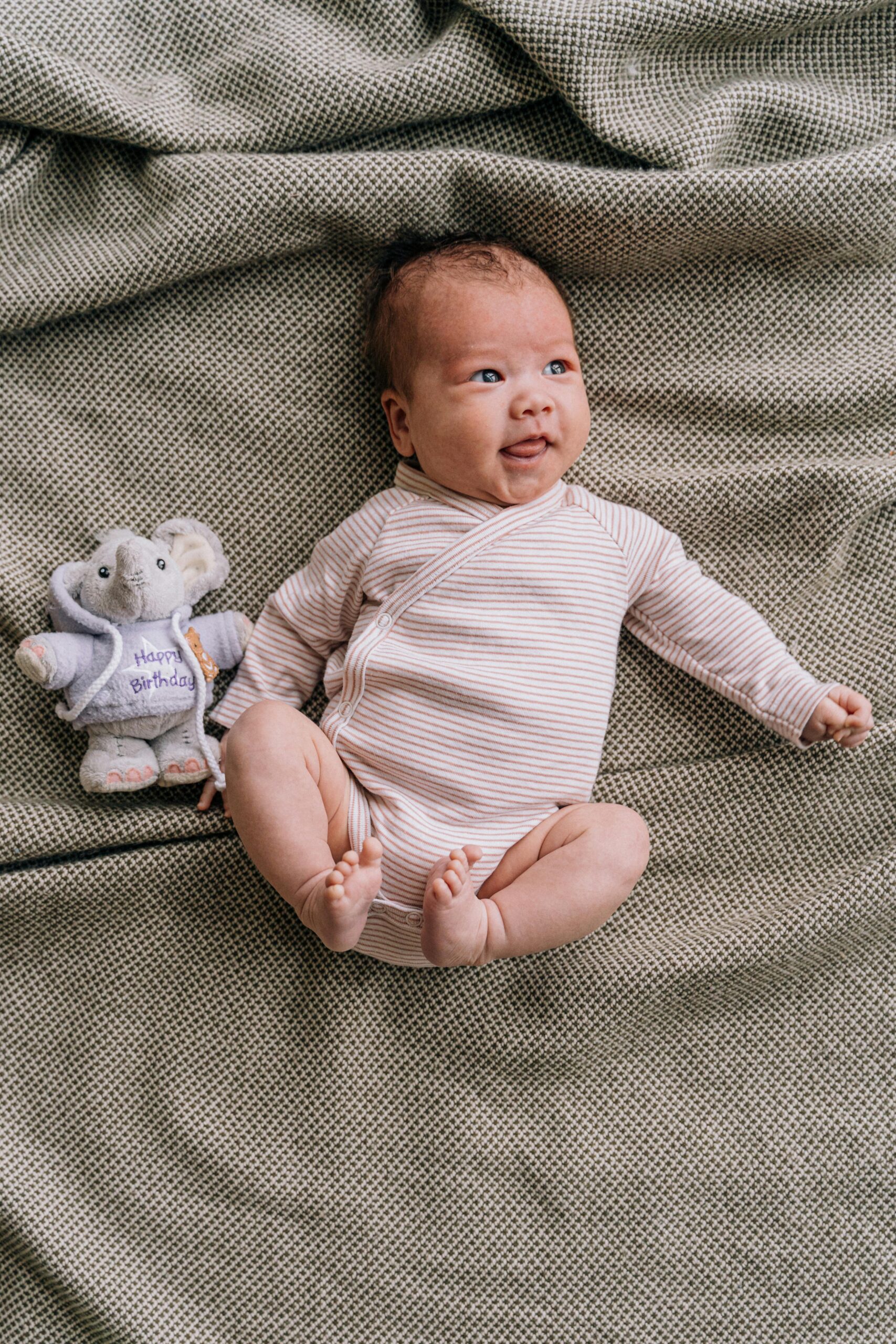 Smiling baby in striped onesie lying on a blanket with a cute stuffed elephant toy.