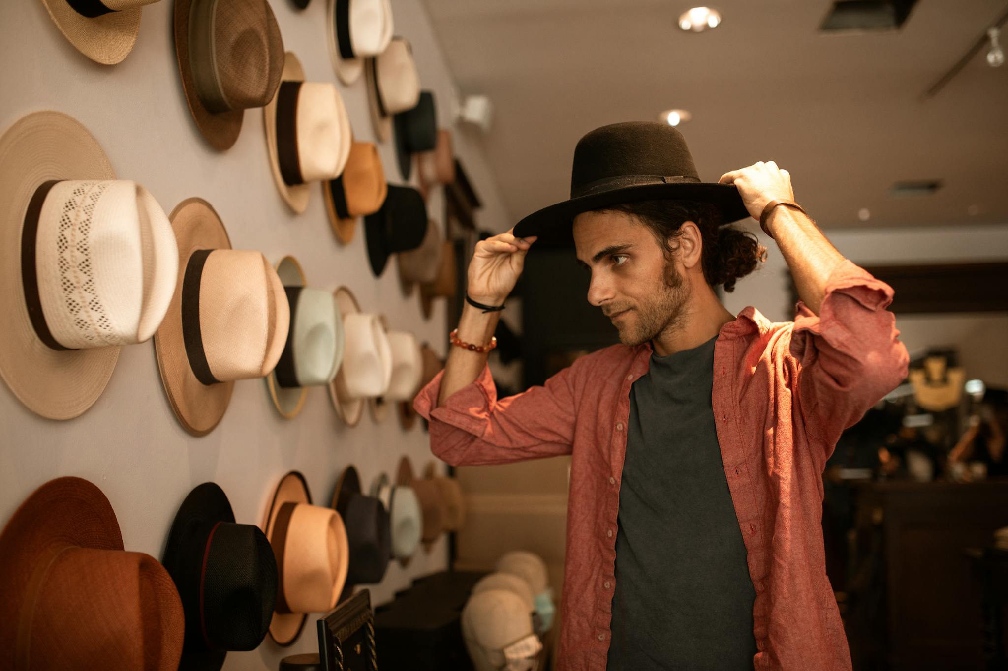 Young man trying on a stylish black fedora in a hat boutique with a varied wall display.